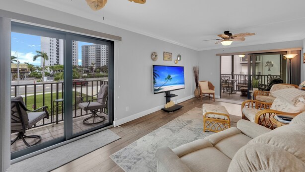 A spacious living room with a large TV, comfortable seating, and a balcony with a view of palm trees and buildings. - 2B/2B Fairview Isles 121 Condo, Walk to Beach, Heated Pool! (Fort Myers Beach)