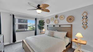 A bedroom with a bed, a ceiling fan, a window with curtains, a bedside table with a lamp, and decorative wall art. - 2B/2B Fairview Isles 121 Condo, Walk to Beach, Heated Pool! (Fort Myers Beach)