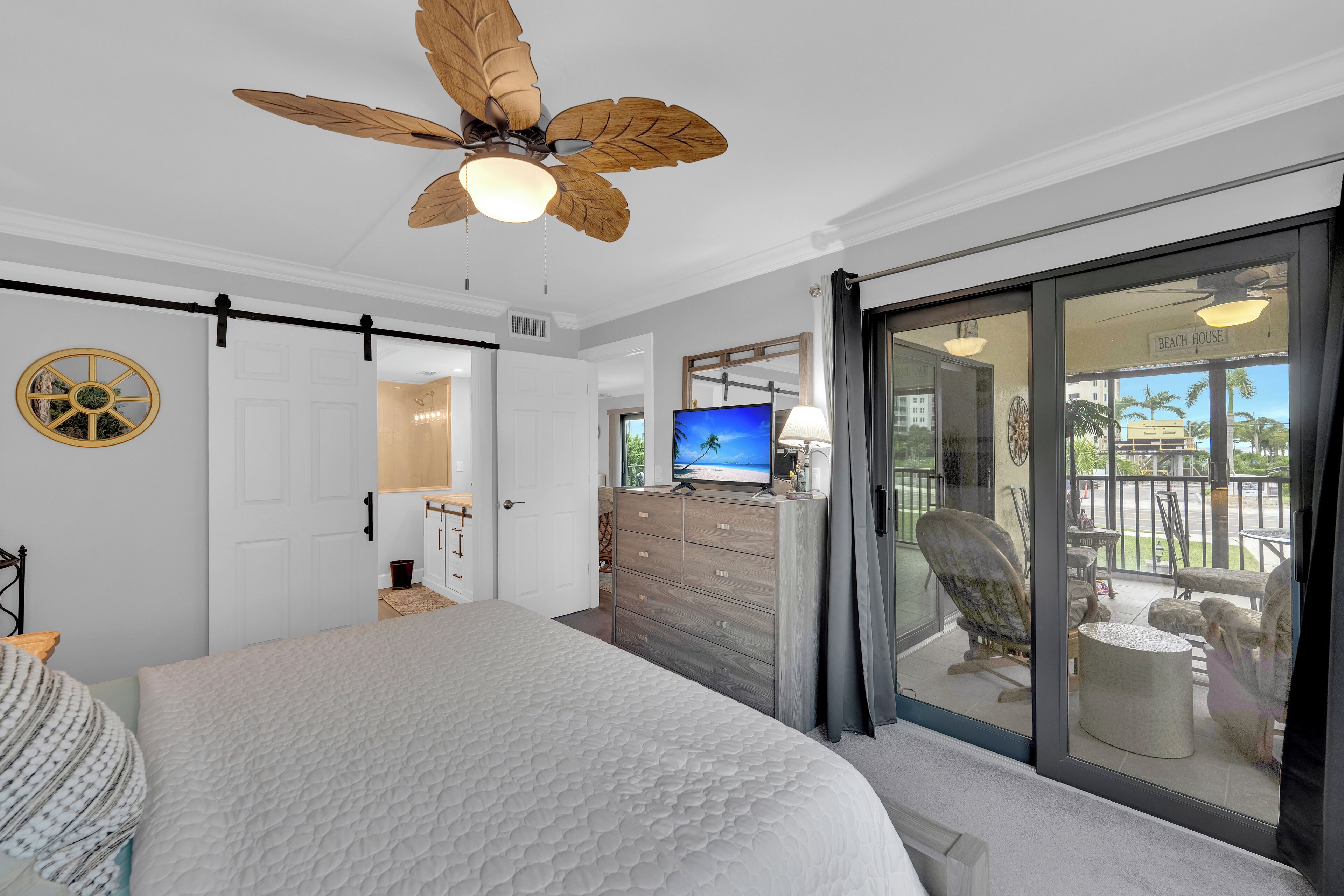 A bedroom with a bed, a ceiling fan, a TV, and a balcony with outdoor furniture.