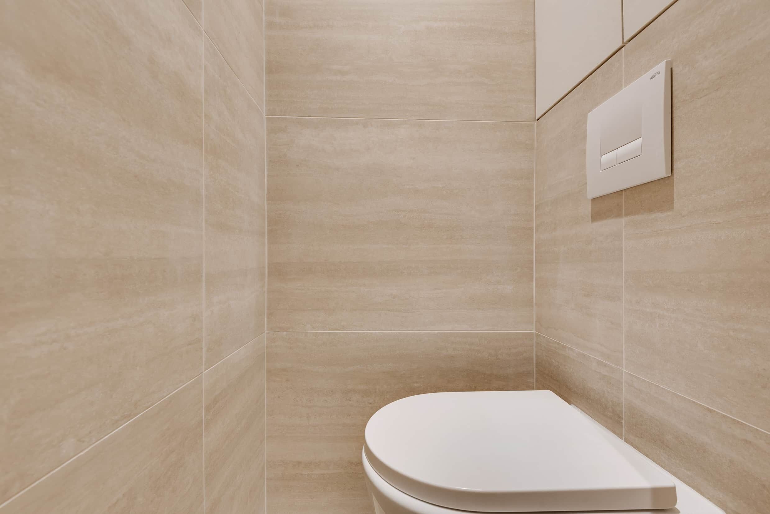 Une salle de bain avec des carreaux de couleur beige, des toilettes blanches et une commande de chasse d’eau fixée au mur.