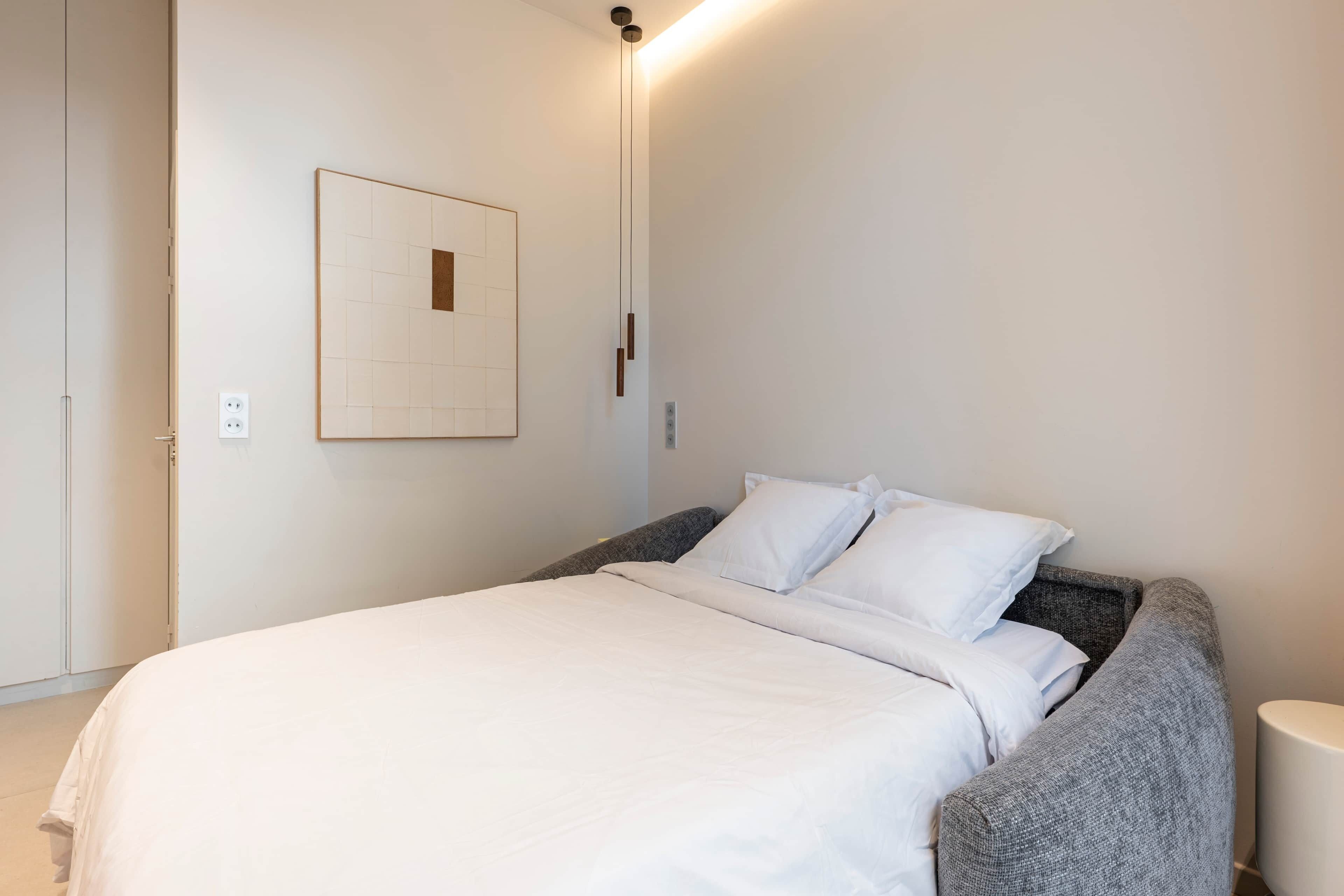 Ein ordentlich bezogenes Bett mit weißer Bettwäsche, einem grau gepolsterten Kopfteil und einer an der Wand montierten Leuchte.
