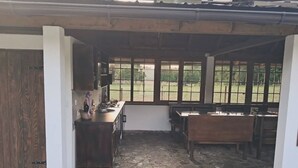Un área de cocina al aire libre techada, con suelo de piedra, mobiliario de madera y grandes ventanales con vistas a un jardín.