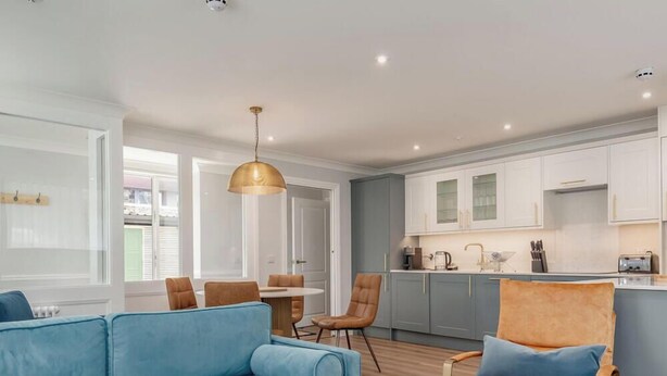A modern living room with a blue sofa, orange chairs, and a kitchen in the background. - Skye Sands | St Mary’s Gardens (No 2) (St Andrews)