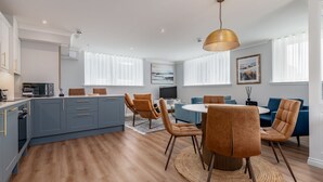 A modern kitchen with a blue island, wooden flooring, and a dining area with brown chairs and a round table. - Skye Sands | St Mary’s Gardens (No 2) (St Andrews)