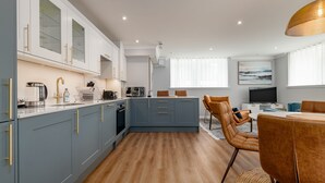 A modern kitchen with blue cabinets, a central island, and a dining area with brown chairs. - Skye Sands | St Mary’s Gardens (No 2) (St Andrews)