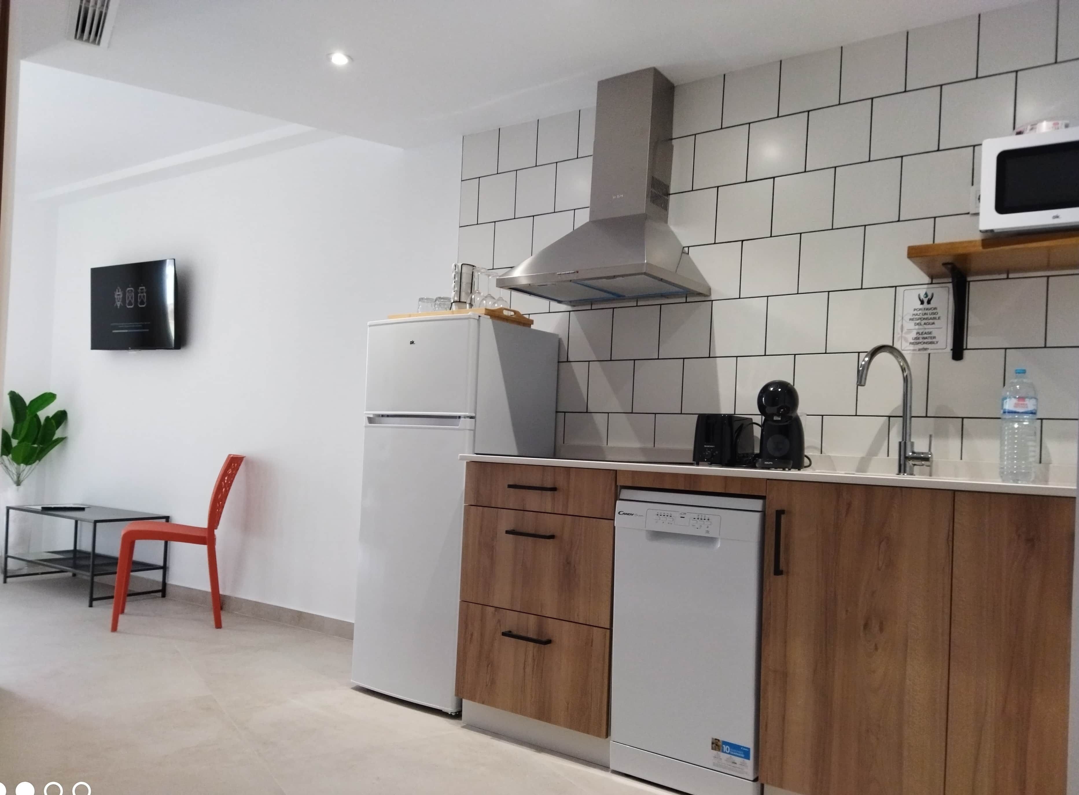 A modern kitchen with a refrigerator, dishwasher, and microwave. There is a wooden cabinet with a sink and a red chair. A potted plant is visible in the corner.