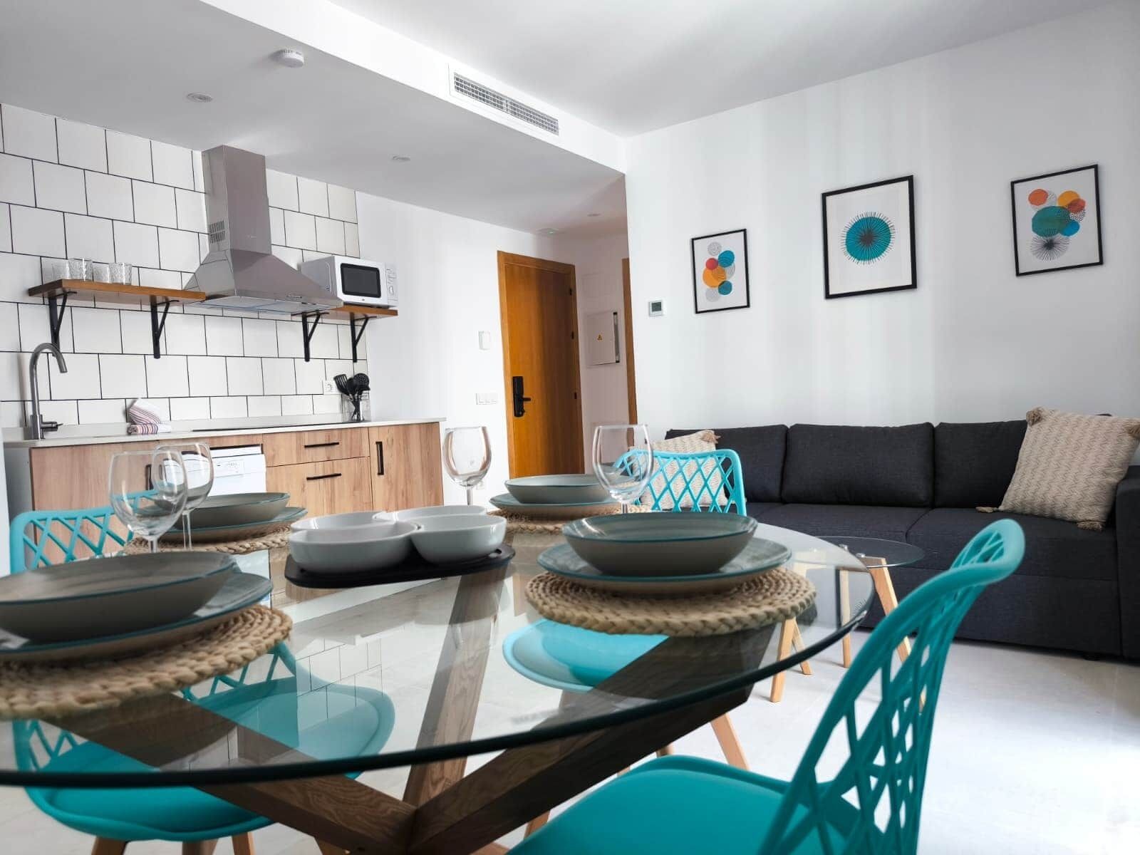 A modern kitchen with white tiles, a built-in microwave, and a stainless steel range hood. There is a dining area with a glass table and turquoise chairs.