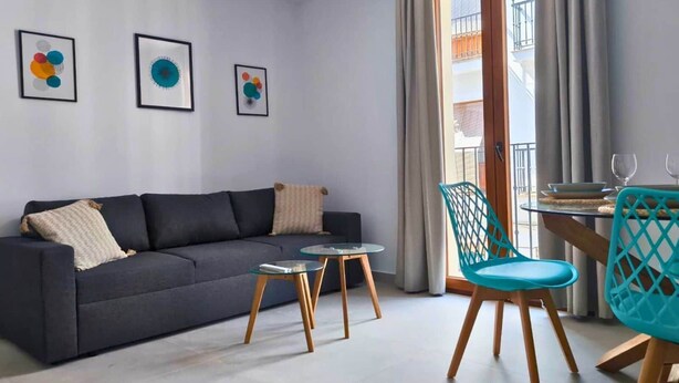 Un salon moderne avec un canapé gris foncé, une table basse en verre et un fauteuil turquoise. Trois œuvres d’art encadrées ornent le mur.