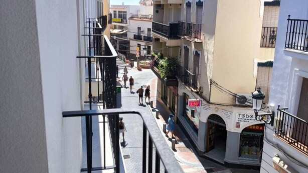 Une rue étroite, bordée de bâtiments des deux côtés, avec un trottoir pour les piétons et une devanture de magasin bien visible, agrémentée d’une enseigne.