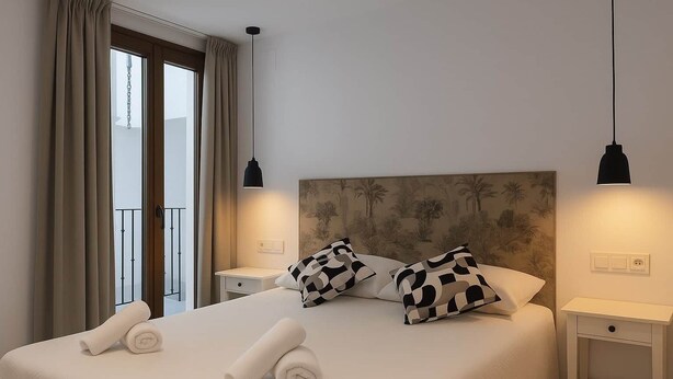 A neatly made bed with rolled towels, two black and white patterned pillows, and a headboard with a palm tree design. There are two bedside tables with black lamps, a window with curtains, and a balcony visible. - Pepa · Serenity Apartment (Nerja)