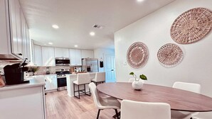 Une cuisine moderne avec des meubles blancs, un coin repas doté d’une table en bois et de chaises blanches, ainsi que des œuvres d’art décoratives sur les murs.