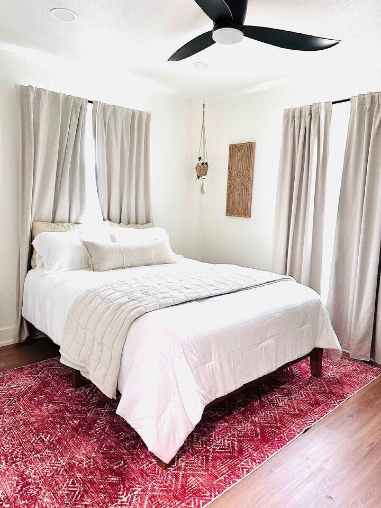 A bedroom with a bed, curtains, a ceiling fan, a wall-mounted plant, and a patterned rug.