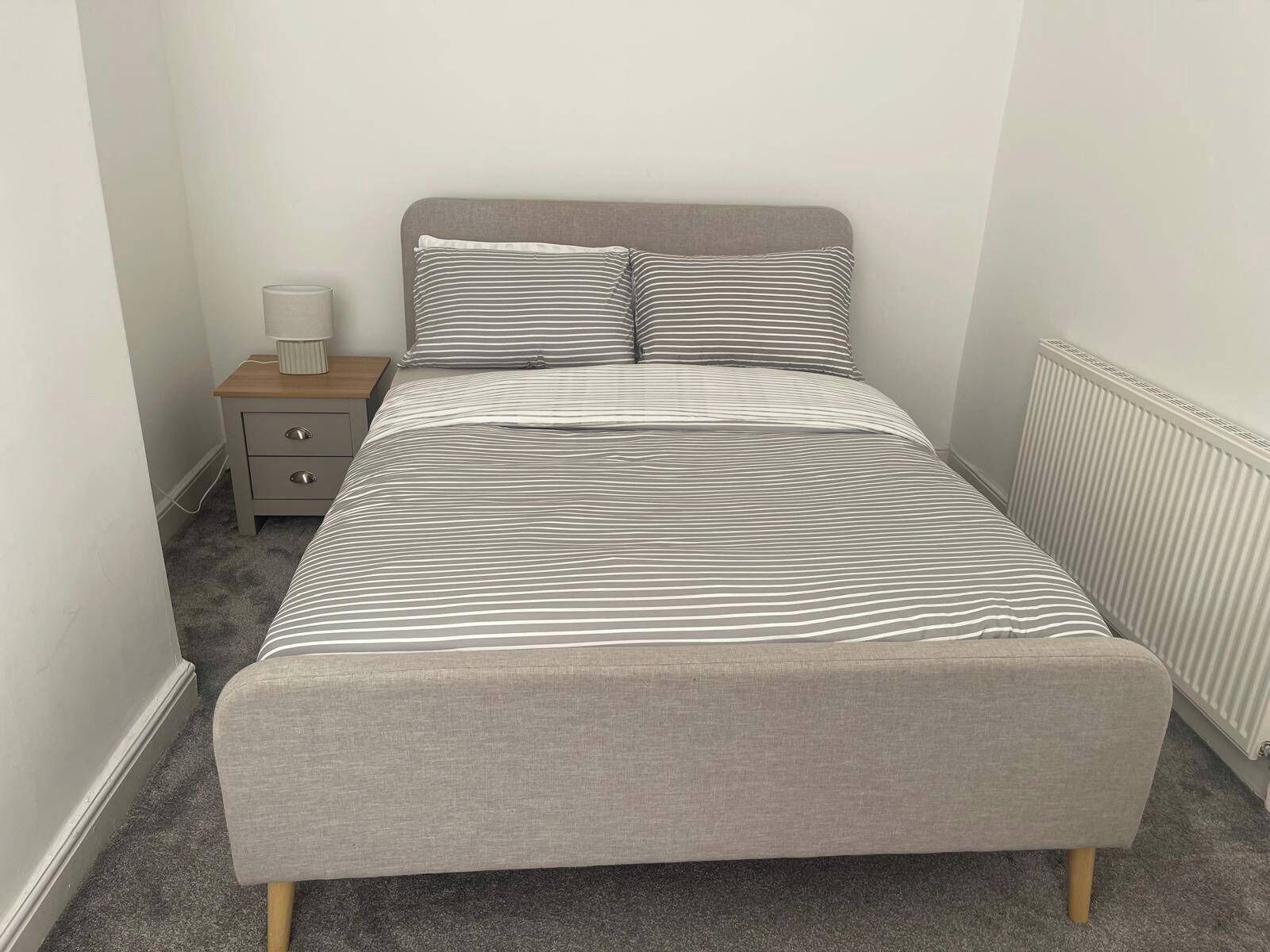 A single bed with a grey headboard and striped bedding in a room with a radiator and a small bedside table with a lamp.