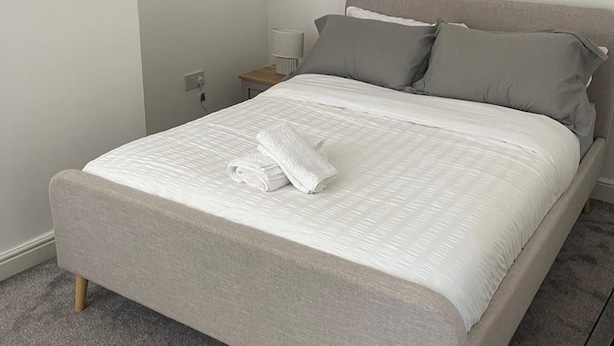 A neatly made bed with a grey headboard and pillows, a rolled white towel, and a bedside table with a lamp. - Manchester Dream House by Cambrian Street (Manchester)