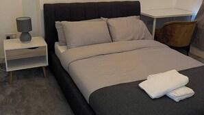 A neatly made bed with a grey and black headboard, a white bedside table with a lamp, and a chair in the corner. - Manchester Dream House by Cambrian Street (Manchester)