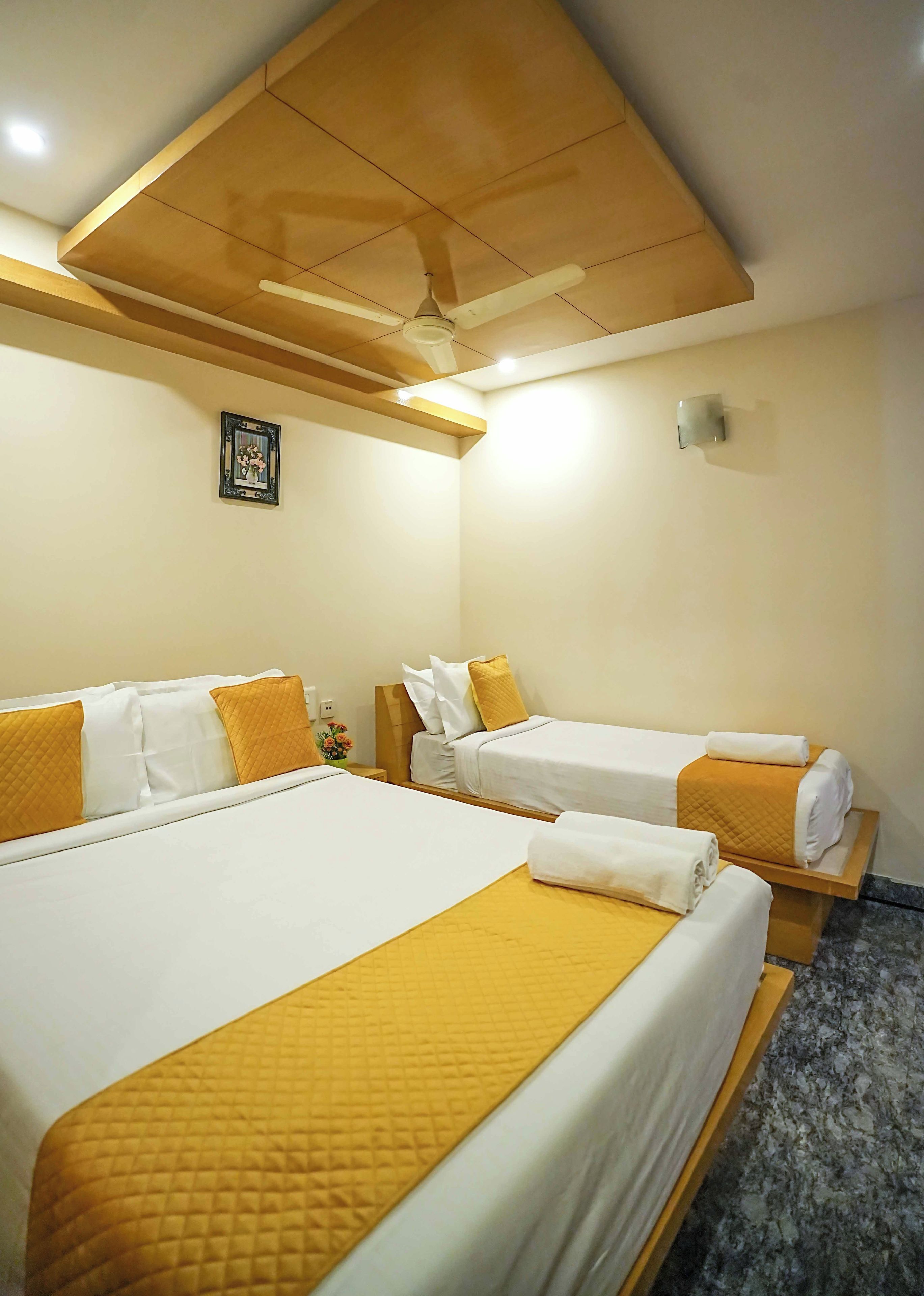 A hotel room with two beds, each with white linens and yellow accents, a ceiling fan, a small framed picture on the wall, and a marble-patterned floor.