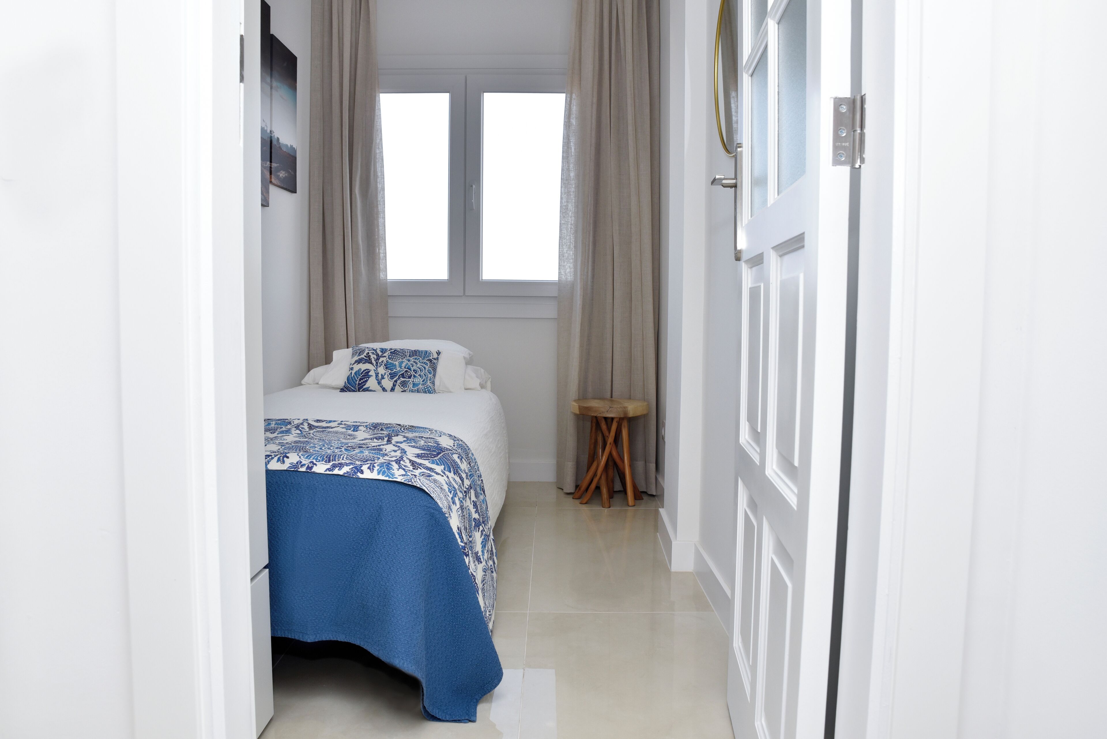 Un dormitori amb un llit, un tamboret petit i una finestra amb cortines.