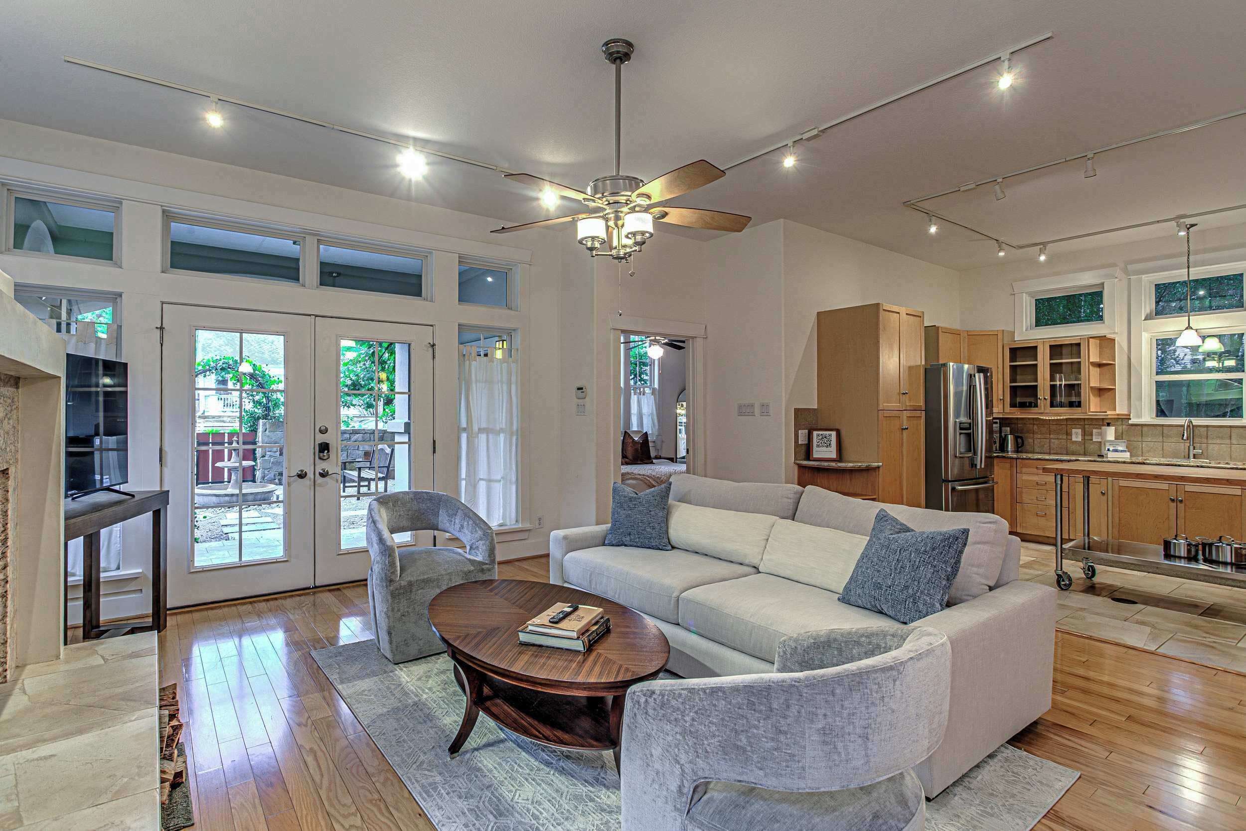A spacious living room with a large sofa, a coffee table, and a ceiling fan. There is a kitchen area with wooden cabinets and a refrigerator.