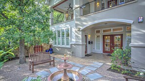 A two-story building with a patio area featuring a stone pathway, a wooden bench, a decorative fountain, and a dogwood tree. - 3BR Retreat near South 1st St. | Walk to SoCo (Austin)
