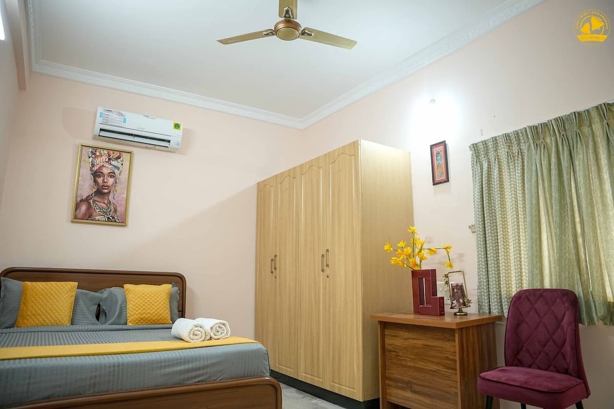 A bedroom with a bed, a wardrobe, a desk with a chair, an air conditioner, a ceiling fan, and a painting on the wall.