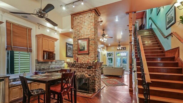A spacious kitchen with a brick fireplace, wooden dining table, and chairs. A staircase with wooden railings leads to an upper level.