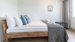 Una cama individual con estructura de madera, ropa de cama blanca y una manta decorativa estampada. Una mesita y un cuadro enmarcado están en la pared.