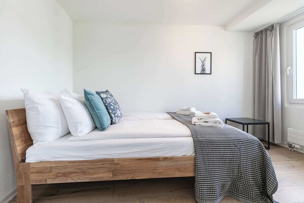 A single bed with a wooden frame, white bedding, and a patterned throw blanket. A small table and a framed picture are on the wall.