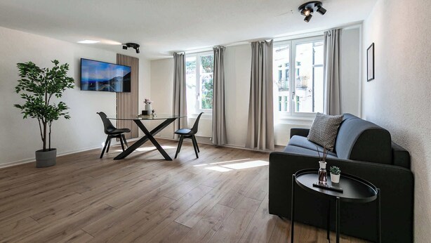 Una sala de estar moderna con una mesa de comedor de vidrio, dos sillas, un sofá, una mesita auxiliar, una planta en maceta, un televisor montado en la pared y un ventilador de techo.