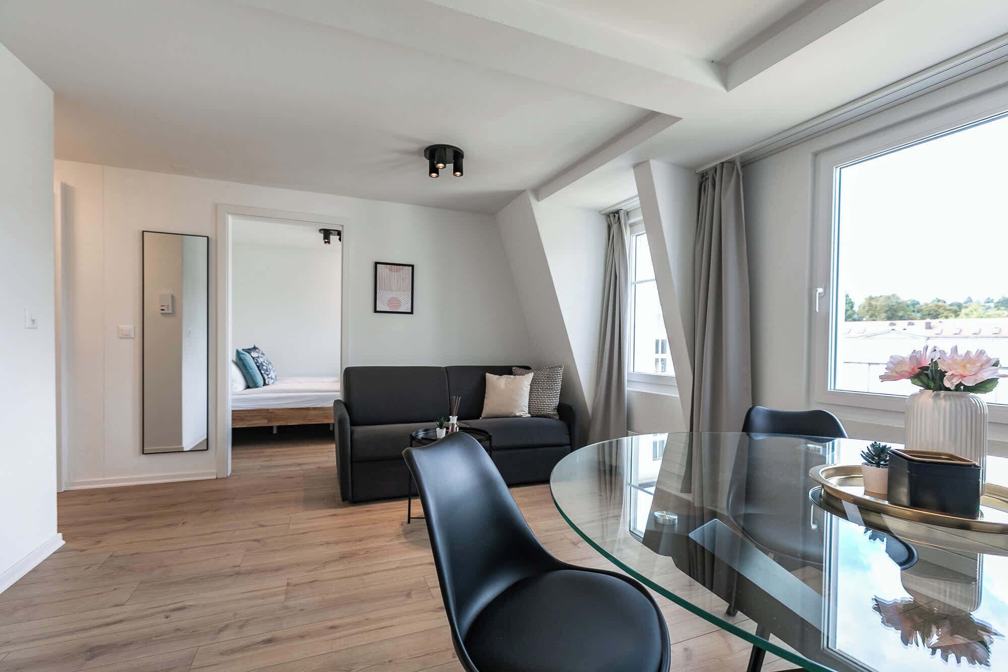 Un salon moderne avec une table à manger en verre, un canapé et un lit à l’arrière-plan.