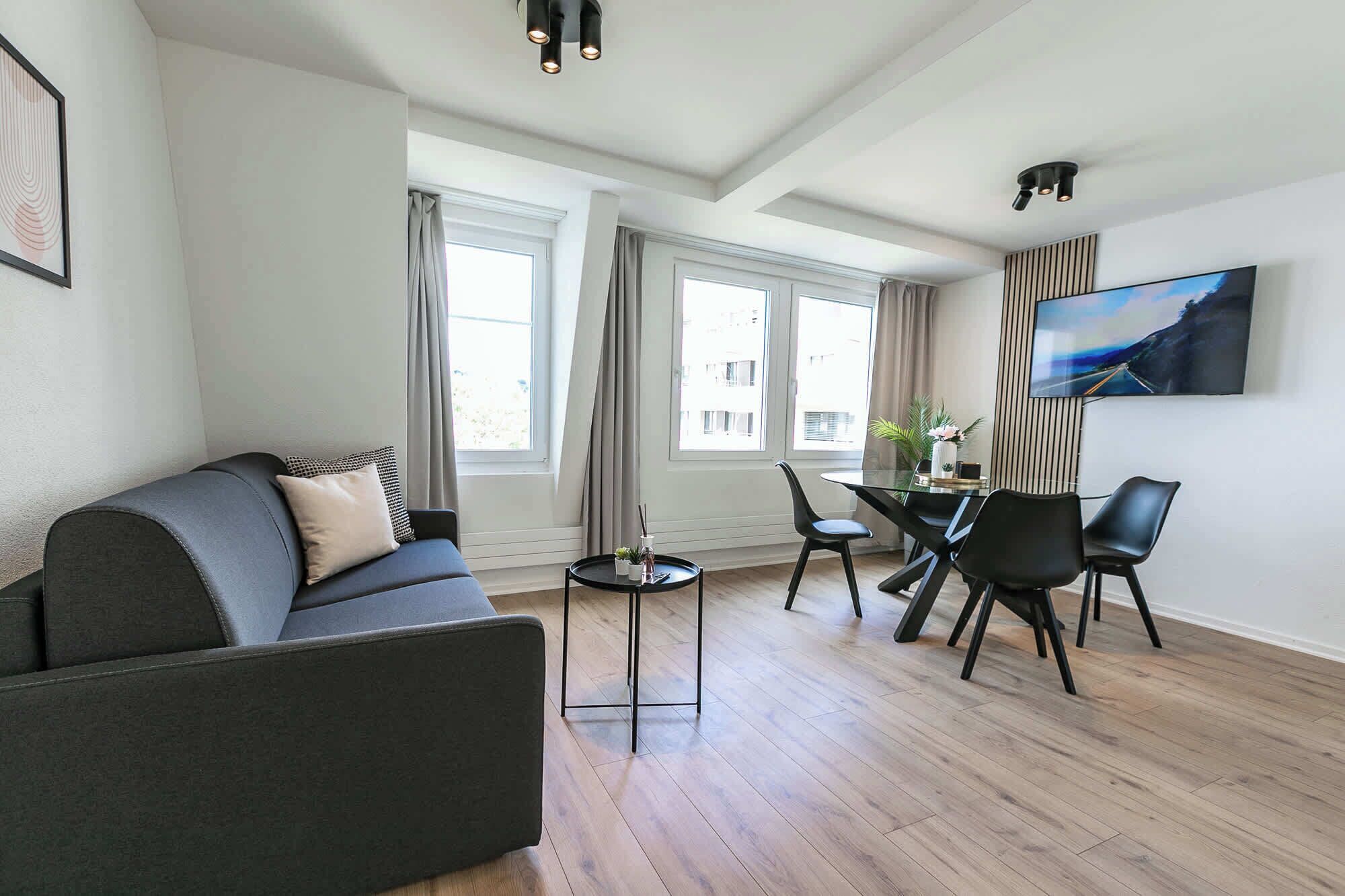 A modern living room with a sofa, dining table, chairs, and a TV mounted on the wall.