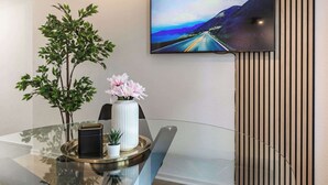 Un intérieur moderne avec une table en verre, une plante en pot, un vase rempli de fleurs, un récipient noir et un téléviseur diffusant une image d’une route pittoresque.
