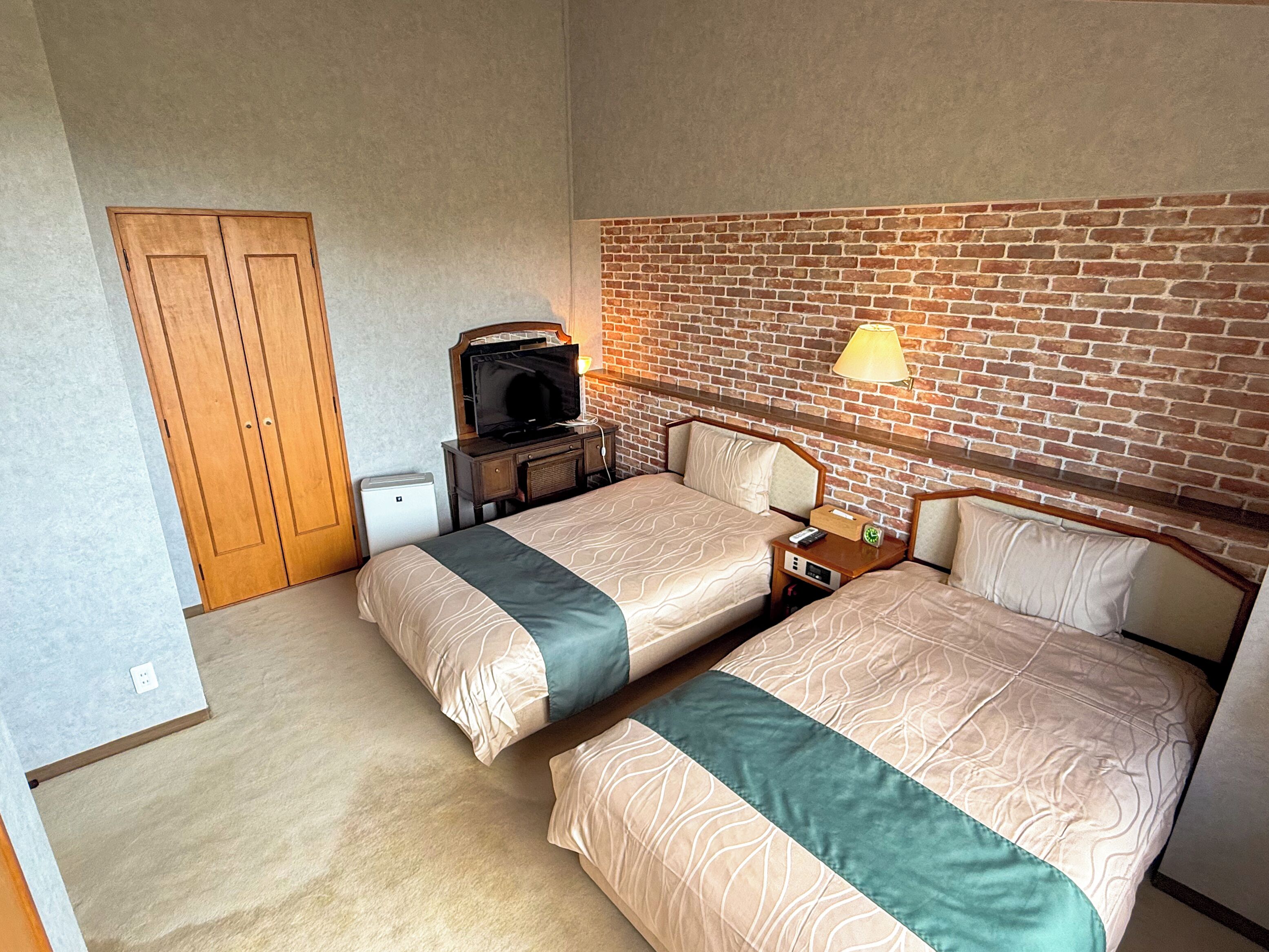 A hotel room with two beds, a television, a lamp, and a brick wall.