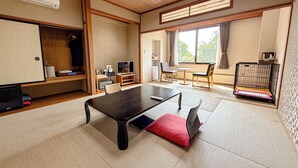 Une pièce avec un sol recouvert de tatamis, une table basse en tatami, une télévision, un petit réfrigérateur et une fenêtre avec des rideaux.