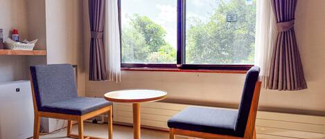 A room with two chairs, a small table, a window with curtains, a radiator, and a shelf with items.