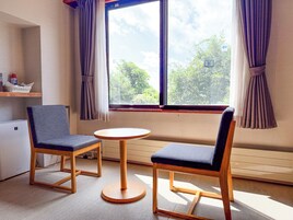 A room with two chairs, a small table, a window with curtains, a radiator, and a shelf with items.
