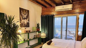 Une chambre avec un lit en bois, une étagère verte ornée de plantes, un tableau, et une vue sur la mer par une grande fenêtre.