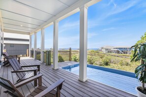 Un espacio al aire libre cubierto con mobiliario de madera en terraza, una piscina y vista al océano y edificios lejanos.