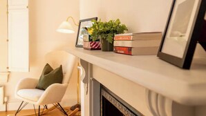 Un salon avec un fauteuil blanc, une petite table avec une lampe, une cheminée et une étagère avec des livres et une plante.