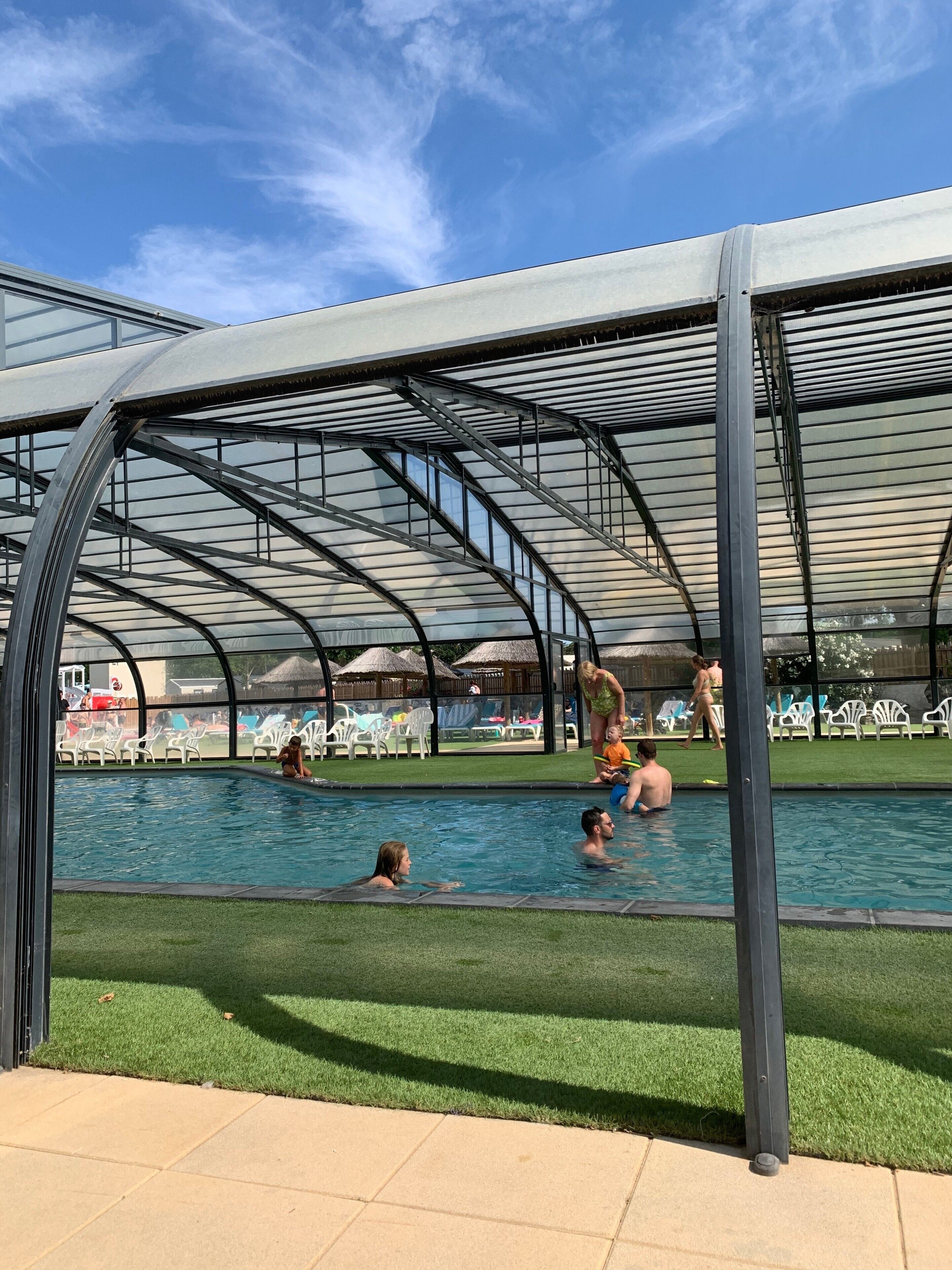 A covered swimming pool with a metal frame and glass panels, surrounded by a grassy area and white plastic chairs.