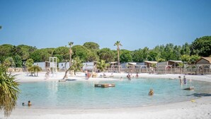 A beach with clear water, palm trees, and small cabins in the background. - Mobile Home at 5* Camping La Yole in Vendres with Lounge Terrace (Vendres)