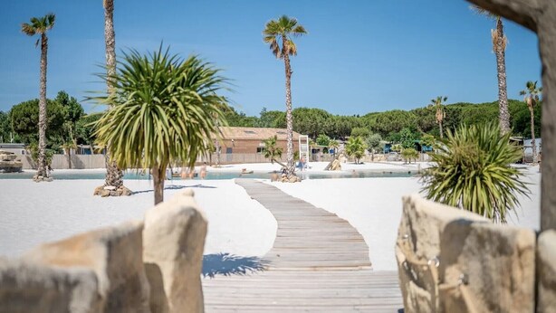 A wooden pathway leading to a beach area with palm trees and a building in the background. - Mobile Home at 5* Camping La Yole in Vendres with Lounge Terrace (Vendres)