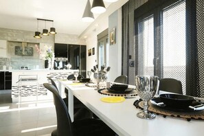 Ein moderner Essbereich mit einem langen Tisch, gedeckt für eine Mahlzeit, einer Küche im Hintergrund und einem großen Fenster mit Moskitonetz.