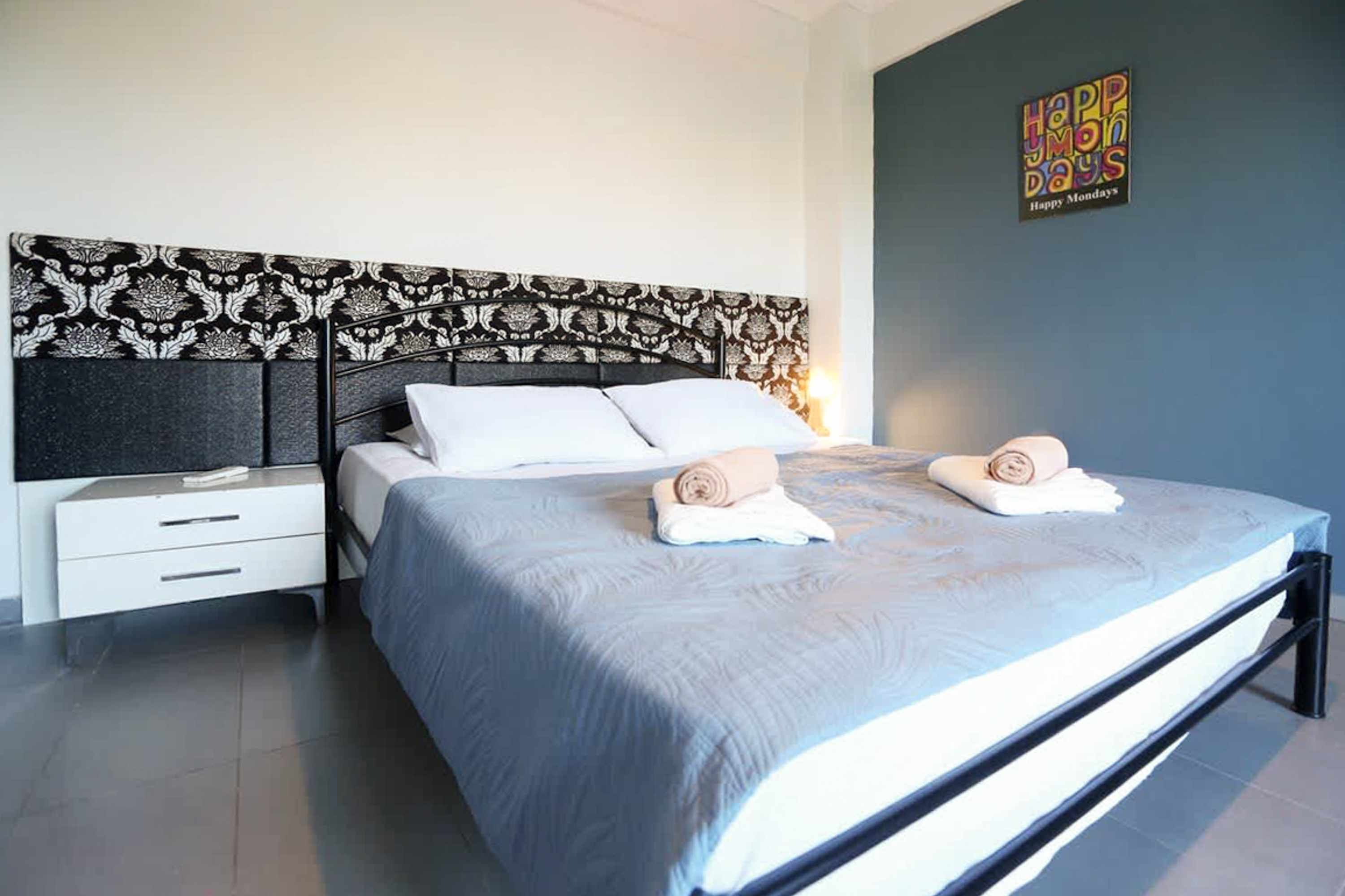 A modern bedroom with a patterned headboard, a neatly made bed with towels, and a 'Happy Monday' sign on the wall.
