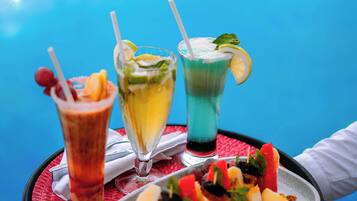 Een dienblad met drie kleurrijke drankjes en een bord met fruitspiesjes, vastgehouden door een persoon.
