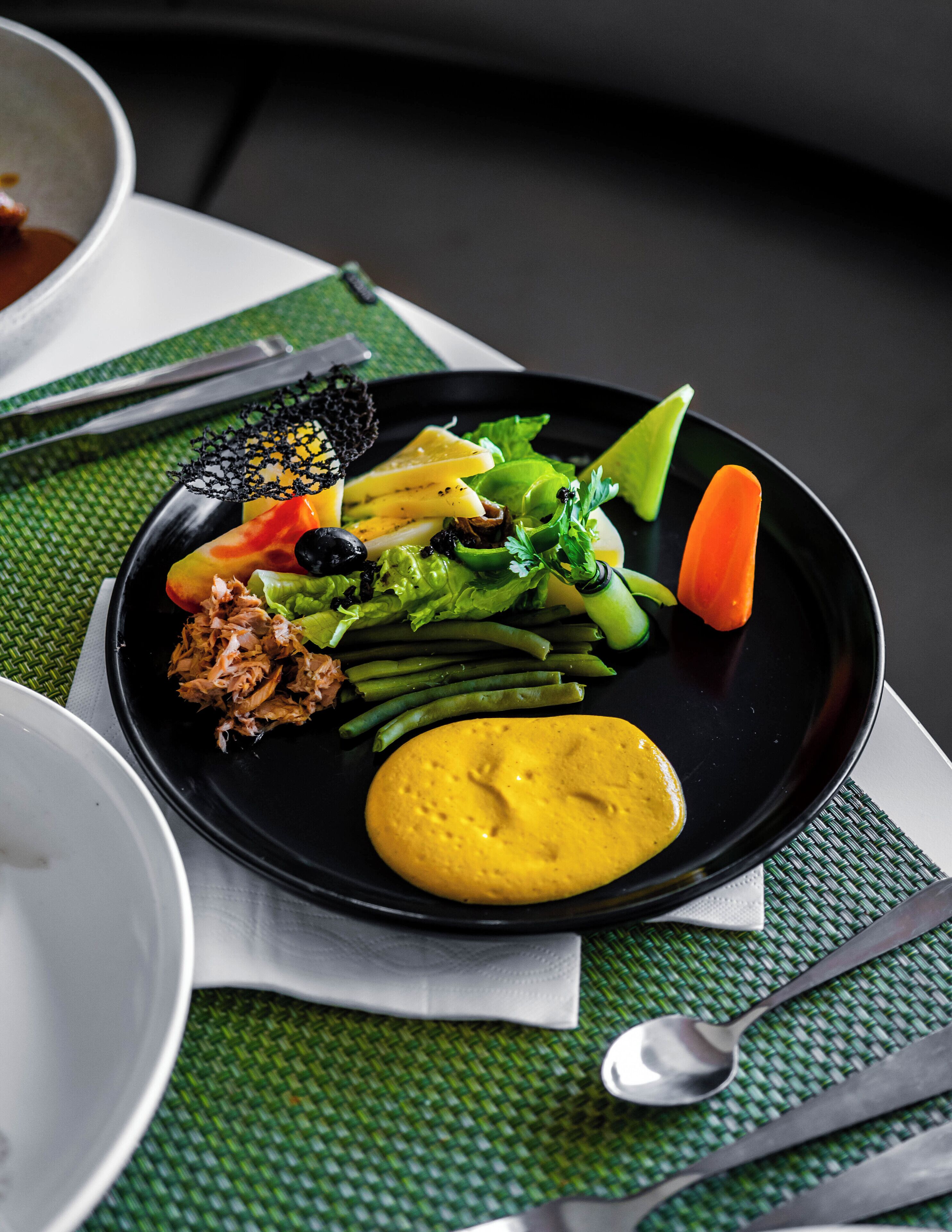 A black plate with various food items including vegetables, a piece of meat, and a yellow sauce, placed on a green woven placemat.