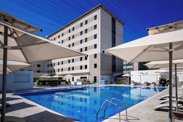 A swimming pool with sun umbrellas, lounge chairs, and a modern building in the background.