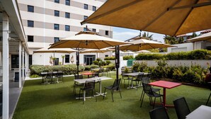 An outdoor patio area with tables and chairs under large umbrellas, surrounded by greenery and a building in the background. - Rénéa Hotel (Sétif)