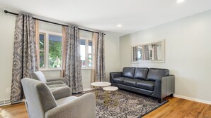 Un salon avec un canapé gris, deux fauteuils gris, un tapis à motifs et une fenêtre avec des rideaux.