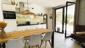Une cuisine moderne avec une table et des chaises en bois, un luminaire suspendu et une vue sur l’extérieur, accessible par une porte vitrée.
