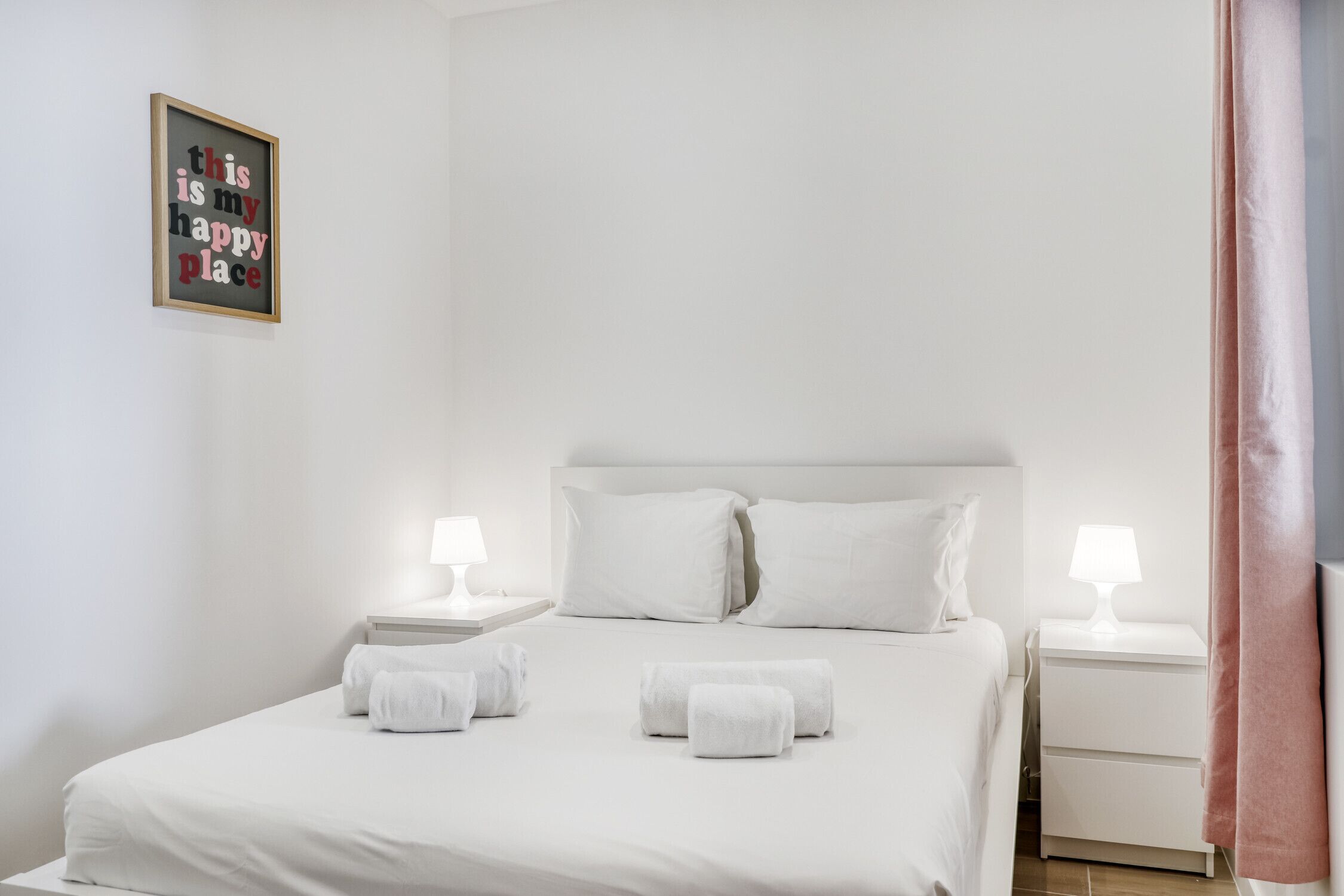 A neatly made bed with white linens and two rolled towels, a bedside table with a lamp, and a framed sign on the wall.